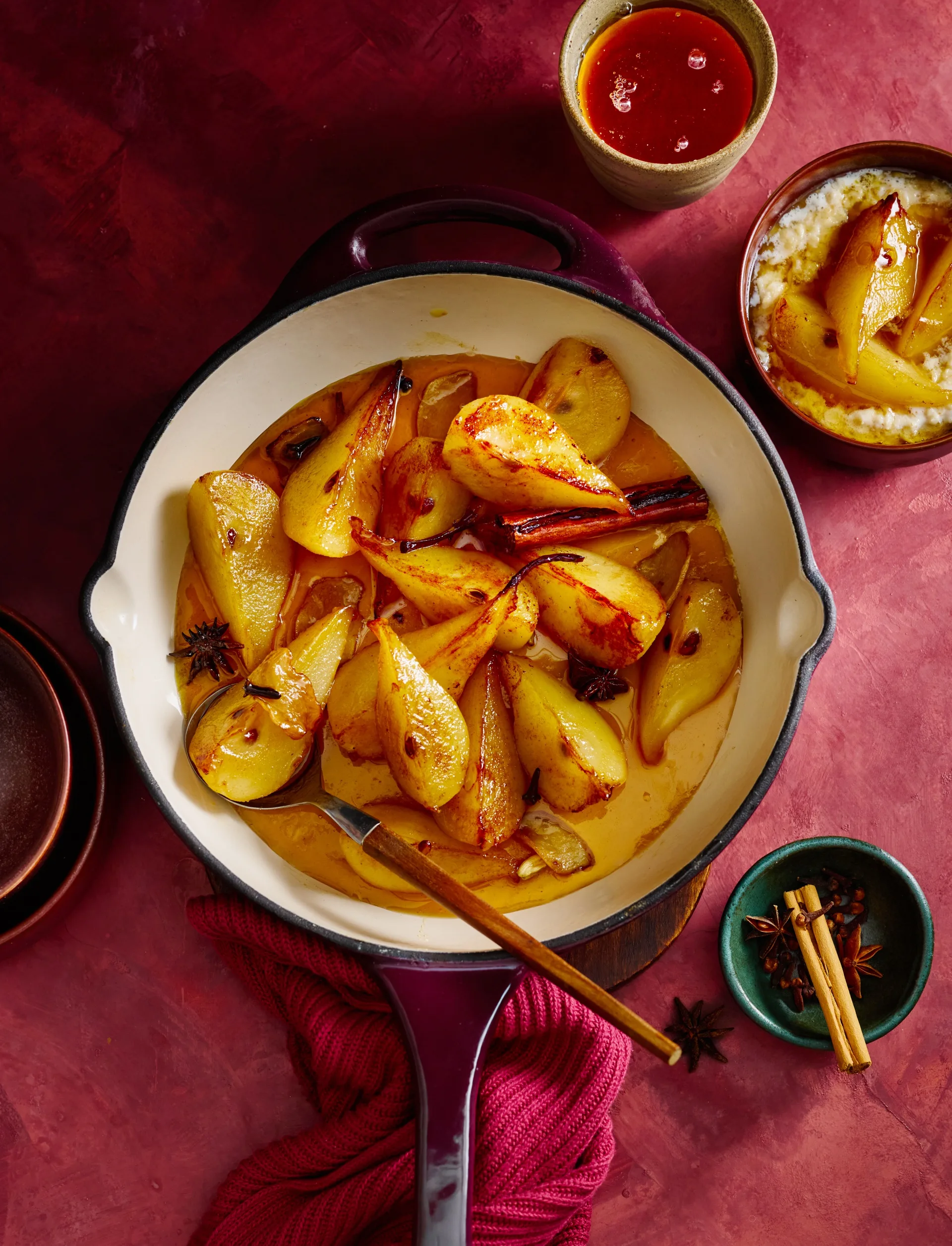 Spiced ginger pears in an enamelled cast-iron pan with a spoon on a red tablecloth.