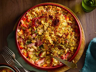 ham hock and white bean bake with breadcrumbs toasted on top in red bowl with spoon sticking out