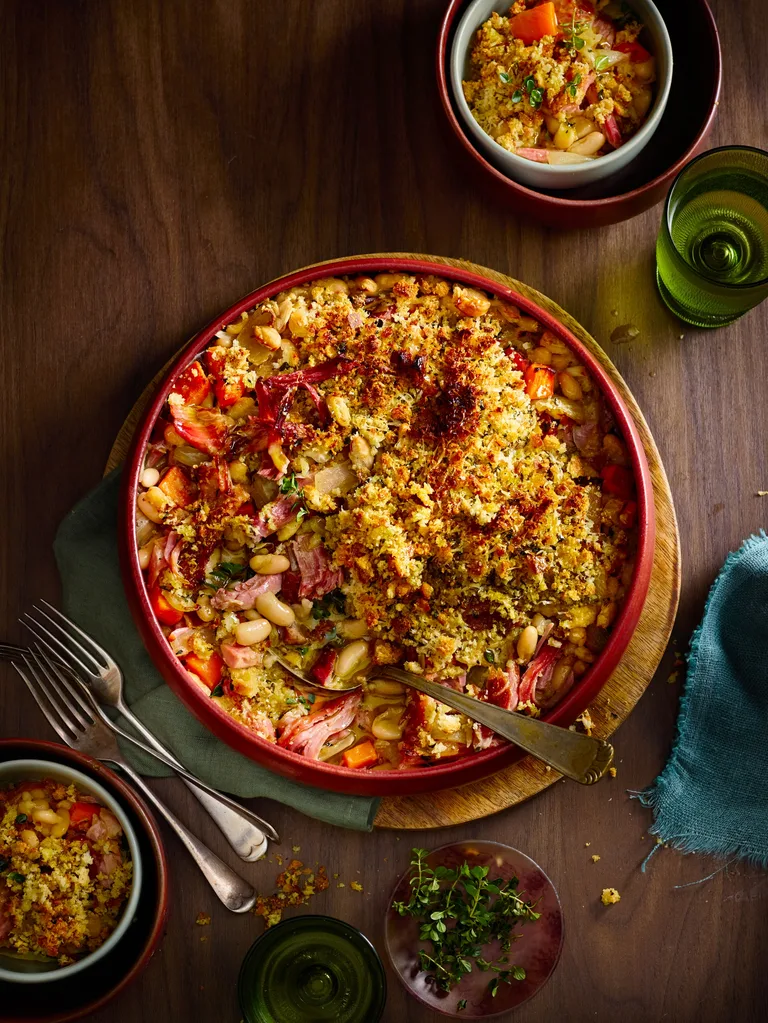 ham hock and white bean bake with breadcrumbs toasted on top in red bowl with spoon sticking out