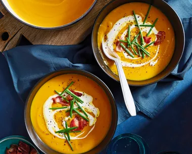two bowls of pumpkin soup with pot in the corner and bowls topped with chives, bacon bits and swirl of cream