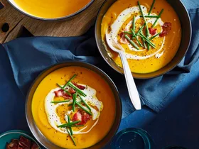 two bowls of pumpkin soup with pot in the corner and bowls topped with chives, bacon bits and swirl of cream
