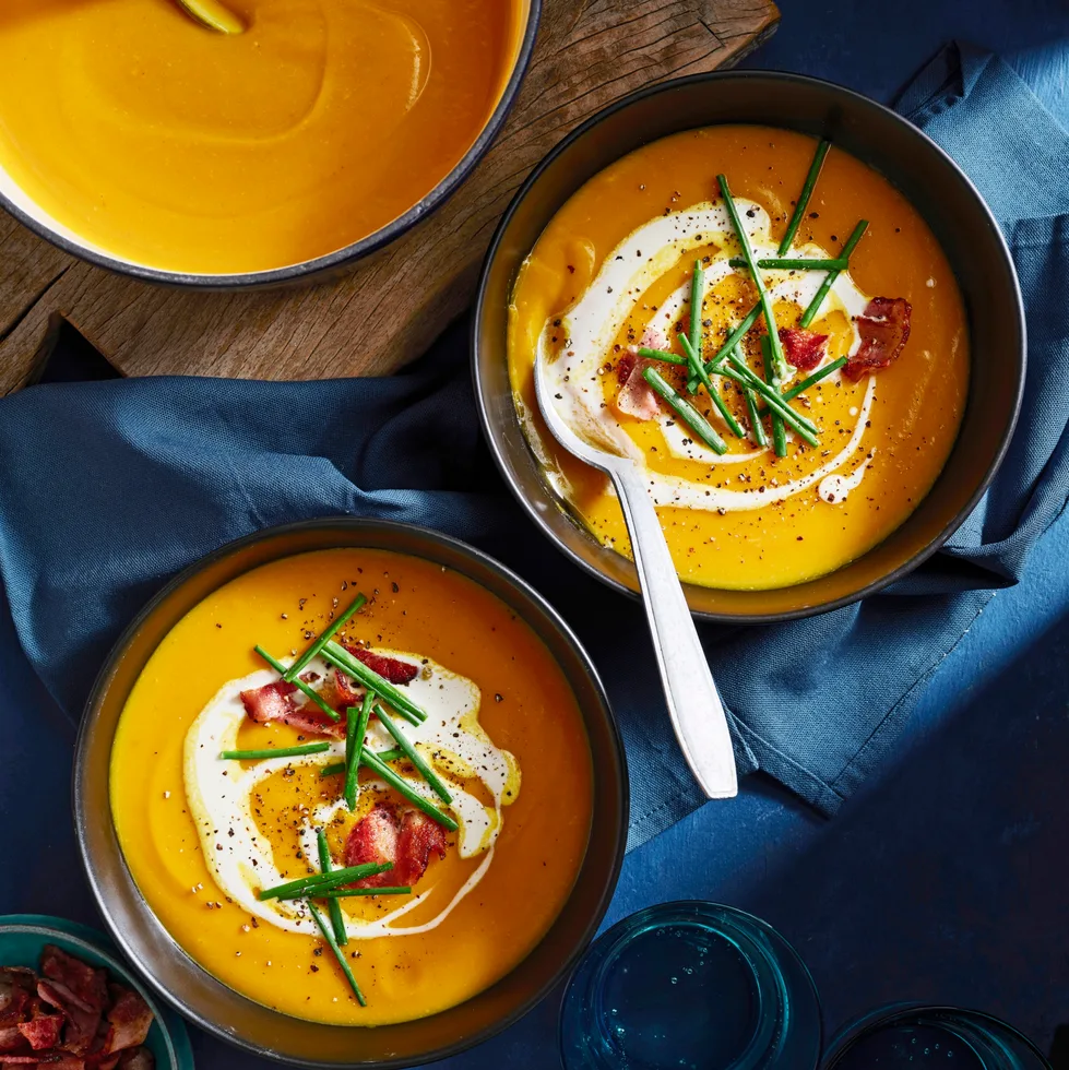 two bowls of pumpkin soup with pot in the corner and bowls topped with chives, bacon bits and swirl of cream