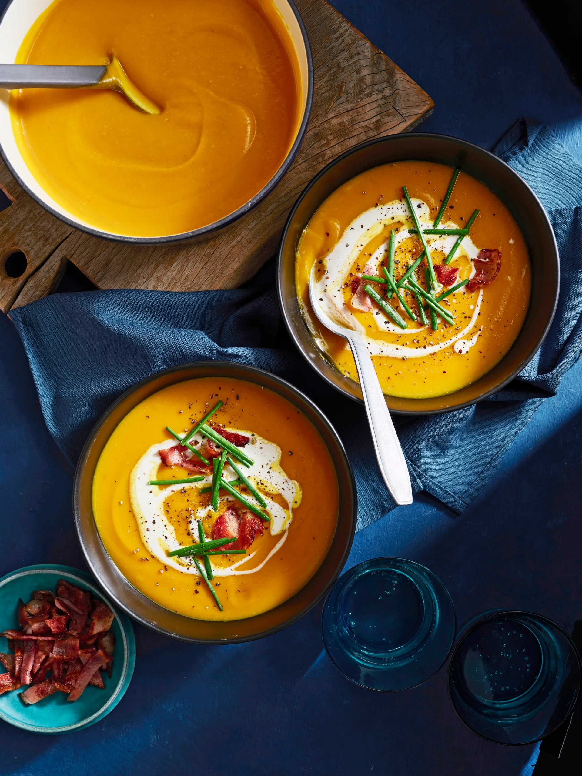 two bowls of pumpkin soup with pot in the corner and bowls topped with chives, bacon bits and swirl of cream