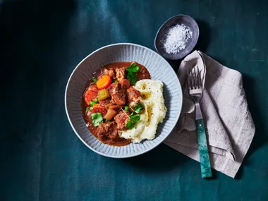 slow cooker beef stew in bowl with side of mashed potato on the bottom and big spoon next to bowl