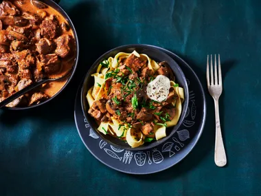 slow cooked beef stroganoff on top of fettucine pasta in grey charcoal bowl with fork on the side