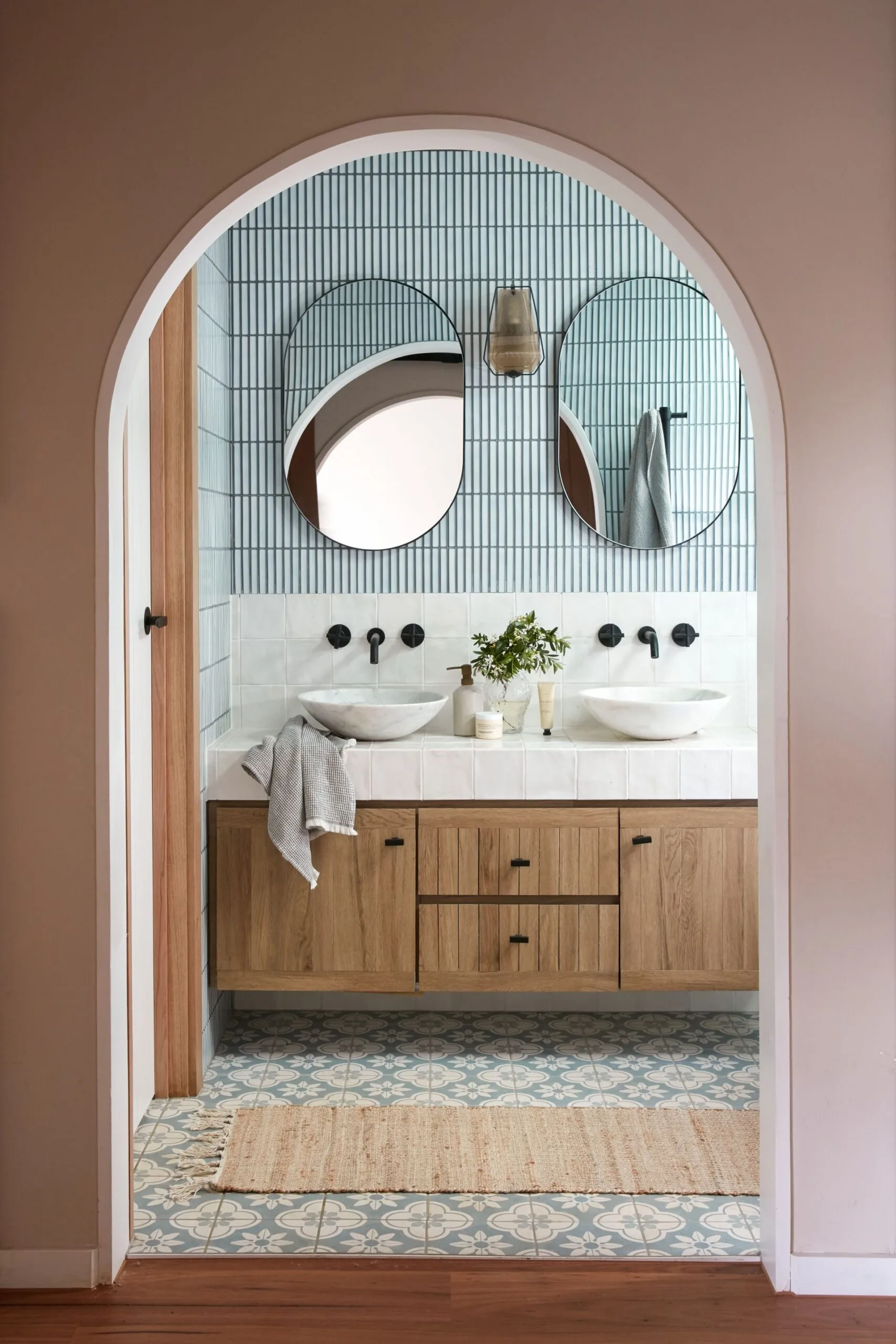 pink painted archway with blue tiled bathroom, two oval mirrors and black tapware above a wooden vanity. 
