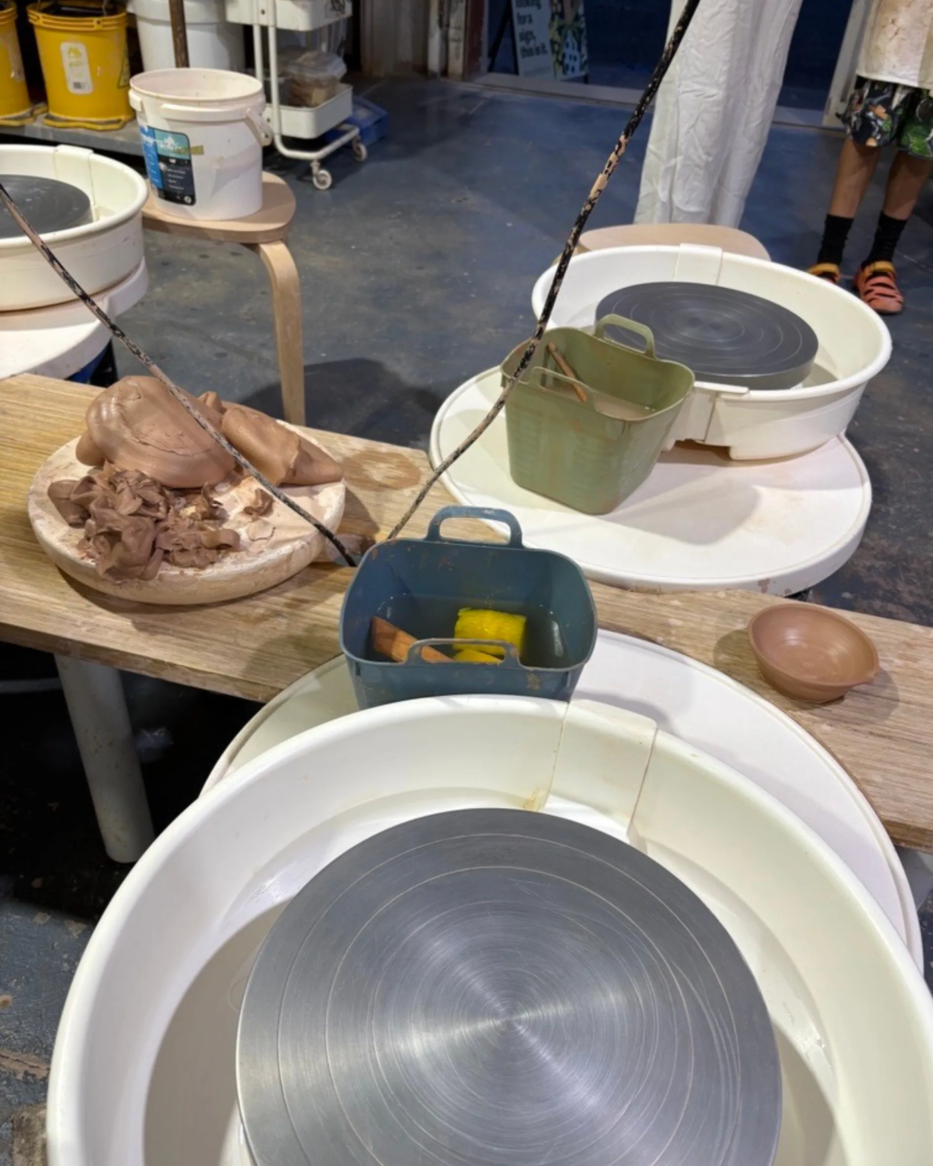 Picture from a pottery class for beginners, a pottery wheel sits in the foreground, on the left, a pile of mangled clay is ready to be destroyed and on the right, there's a bowl