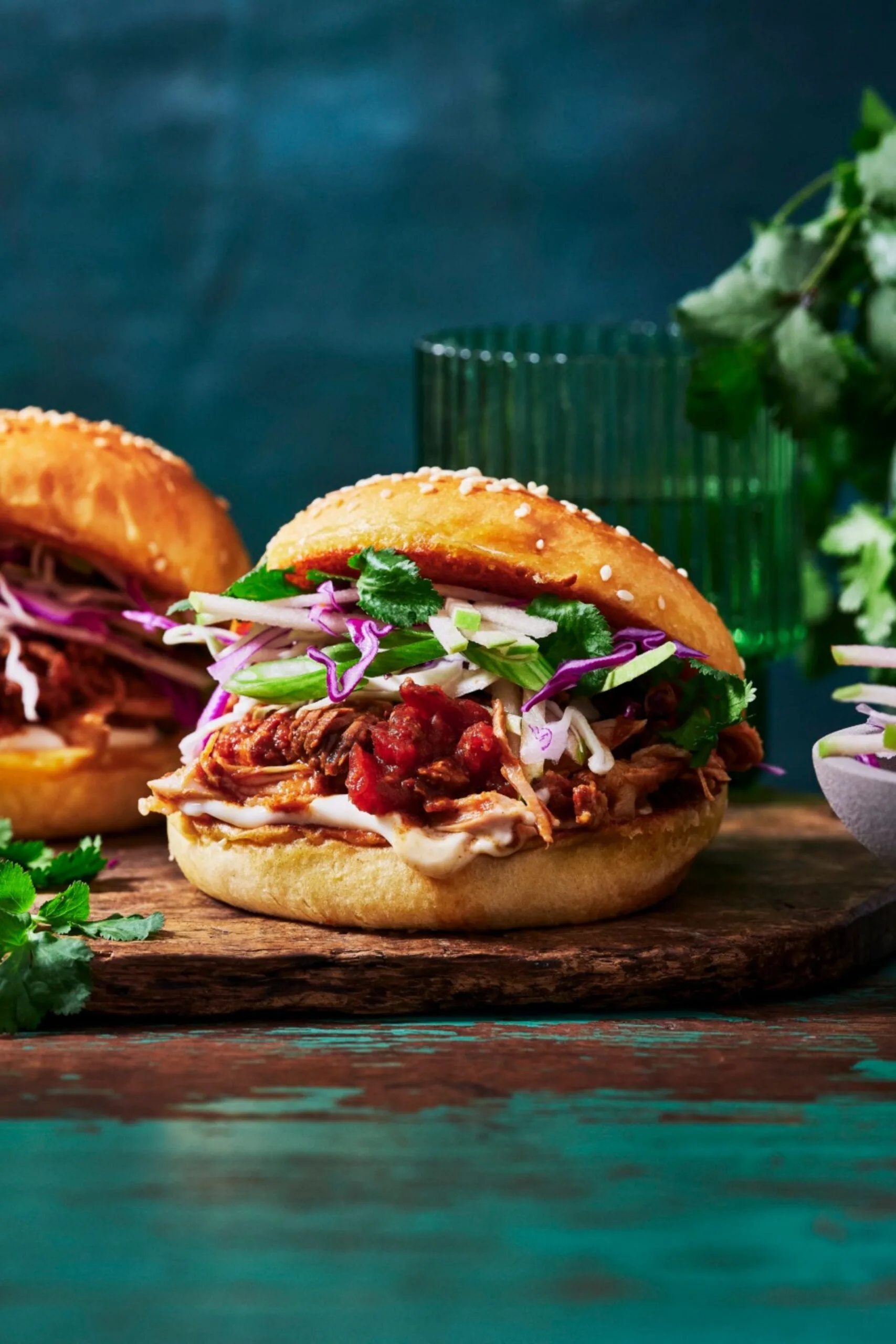 close up of pulled pork bruger with bbq sauce on the bottom, cabbage slaw and a potato bun all on a wooden chopping board