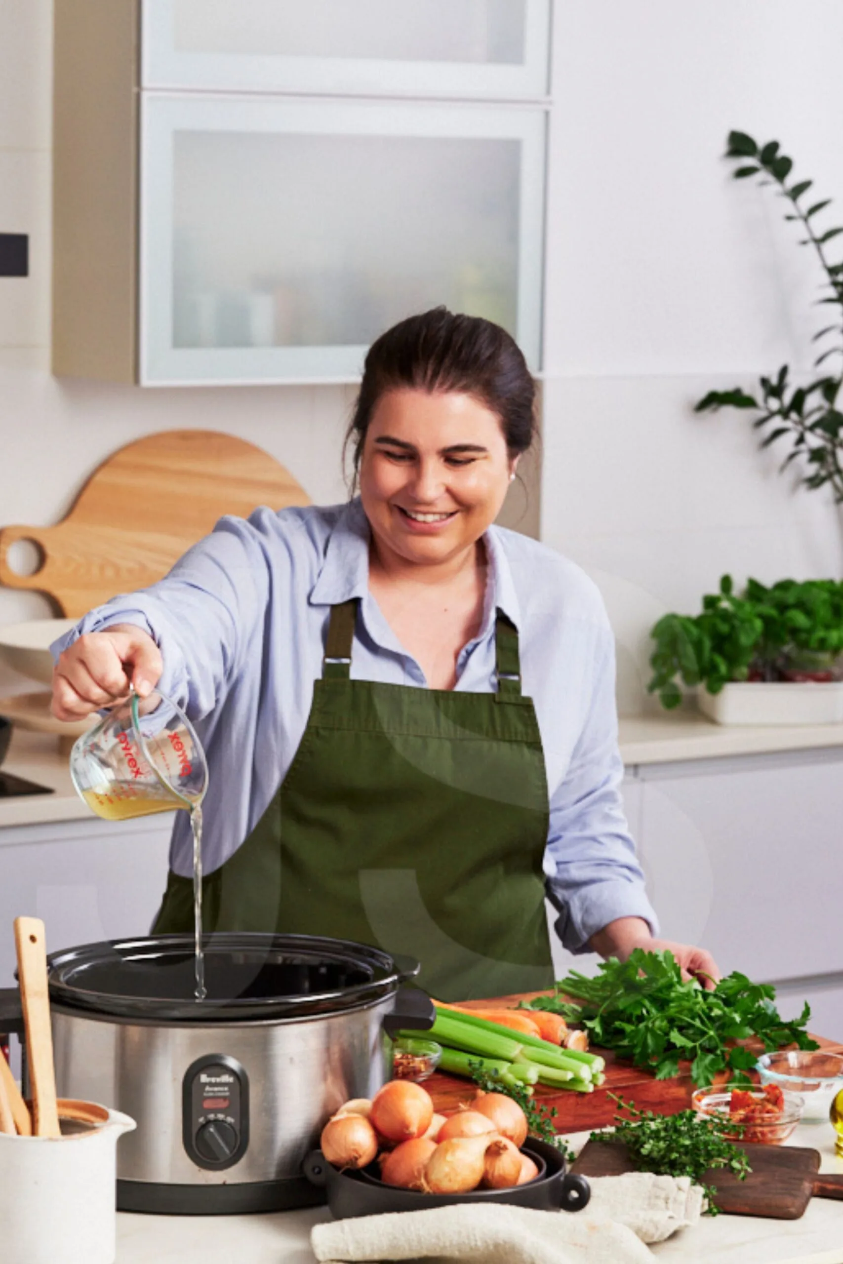 Sarah Murphy pouring stock into the slow cooker with a tray of vegetables including celery carrot and onion on chopping board in front of her