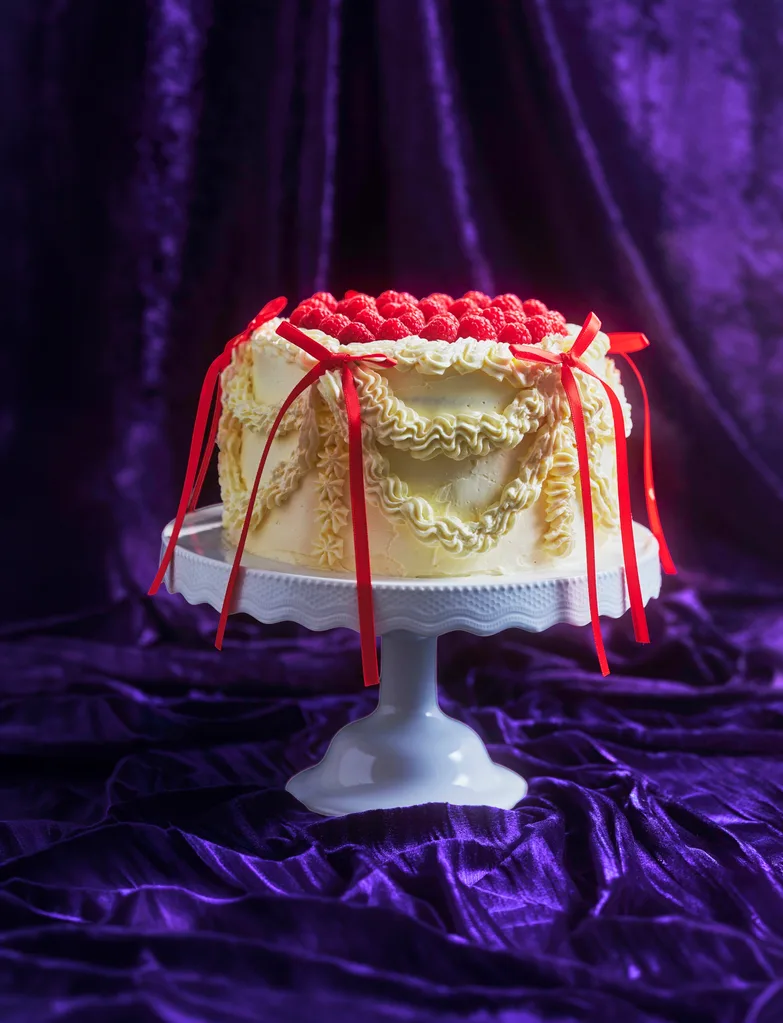white cake with rasberrys on top and red ribbons on a white cake stand with a purple velvet backdrop