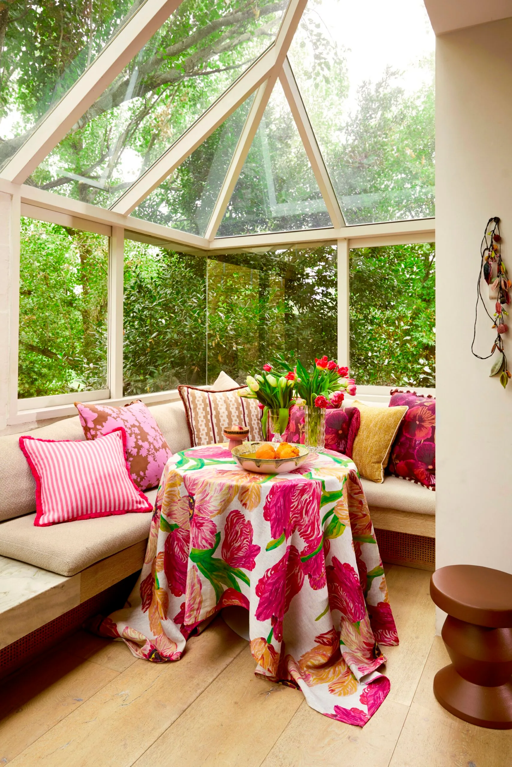 Glass sunroom with banquette seating, pink cushions, a vase of tulips and a floral tablecloth