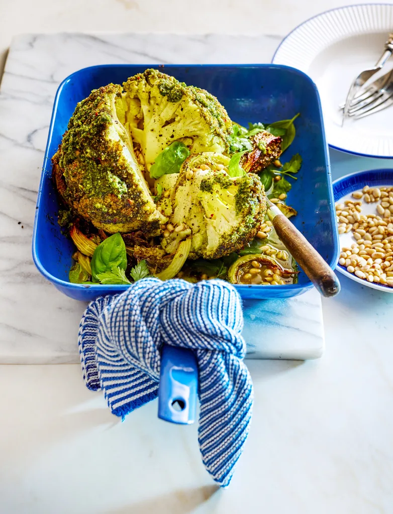 whole baked cauliflower with herby green butter on top, on a blue roasting dish with tea towel on handle