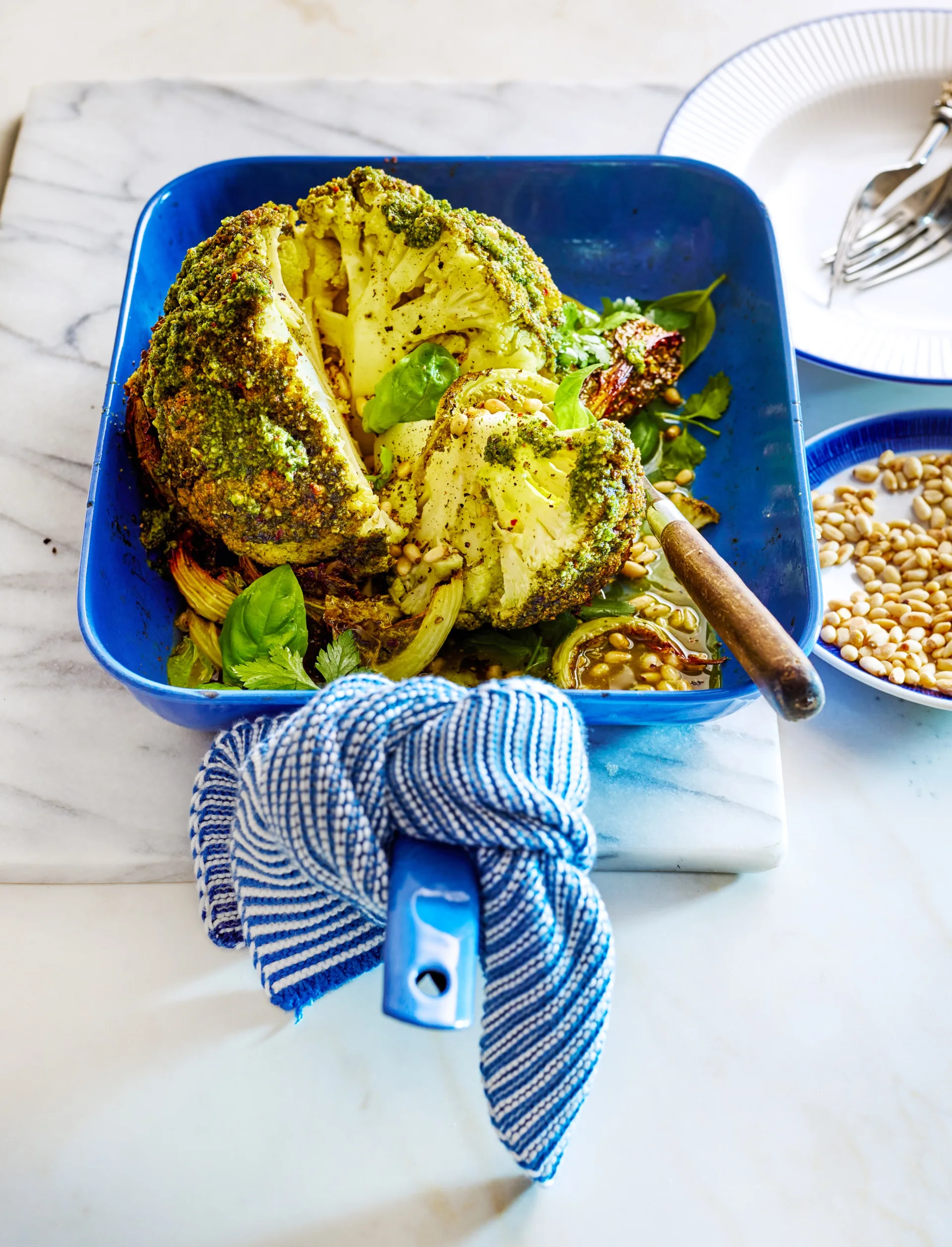 whole baked cauliflower with herby green butter on top, on a blue roasting dish with tea towel on handle