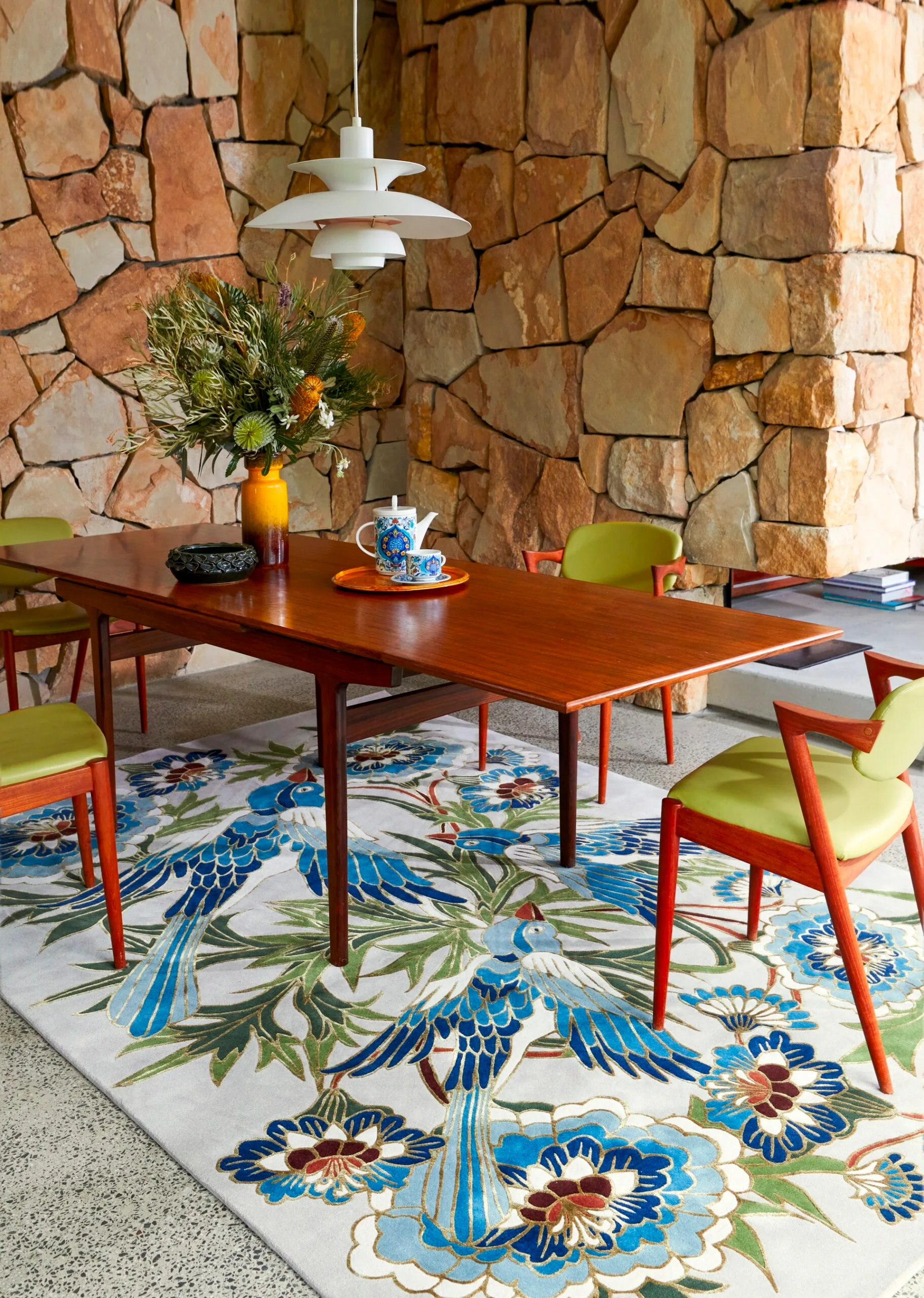 Midcentury Parker style dining table and green chairs in front of a stone wall with a floral rug