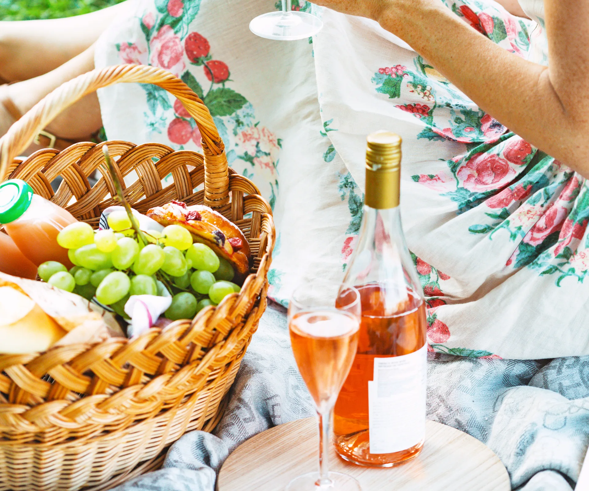 picnic set up with wicker basket filled with grapes, bread and cheese. Sitting on cheeseboard is rose bottle and filled glass