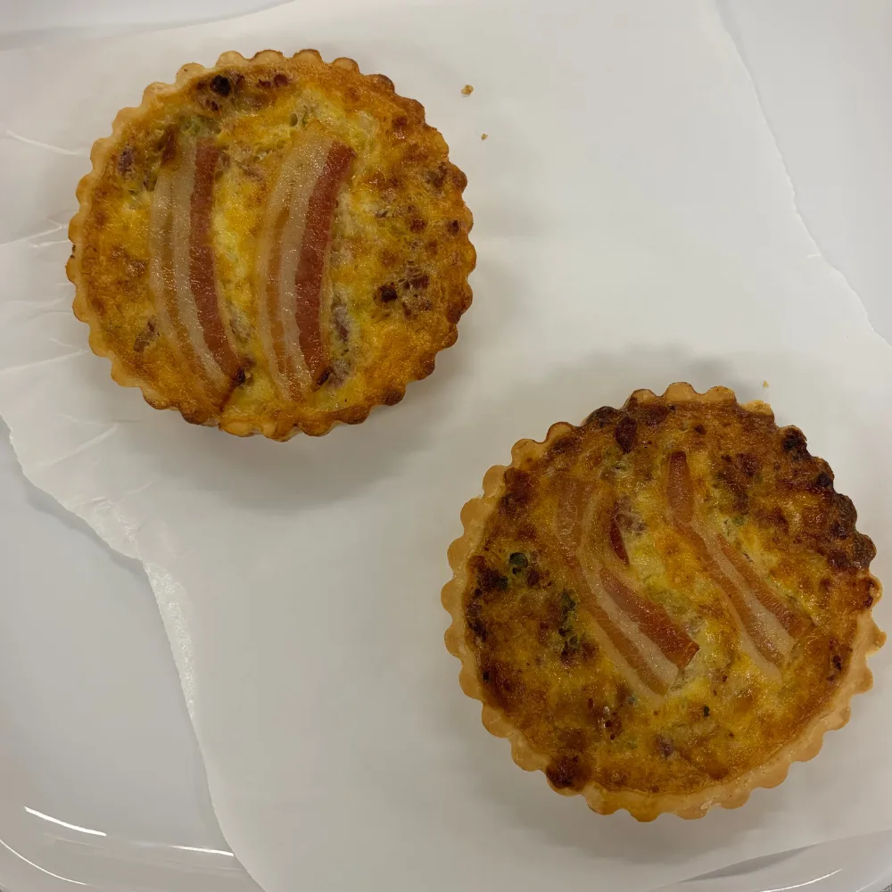 Signature Collection Double Smoked Bacon & Gruyere Quiche with Sauteed Leek two quiches with two pieces of bacon on each on top sitting on white tray