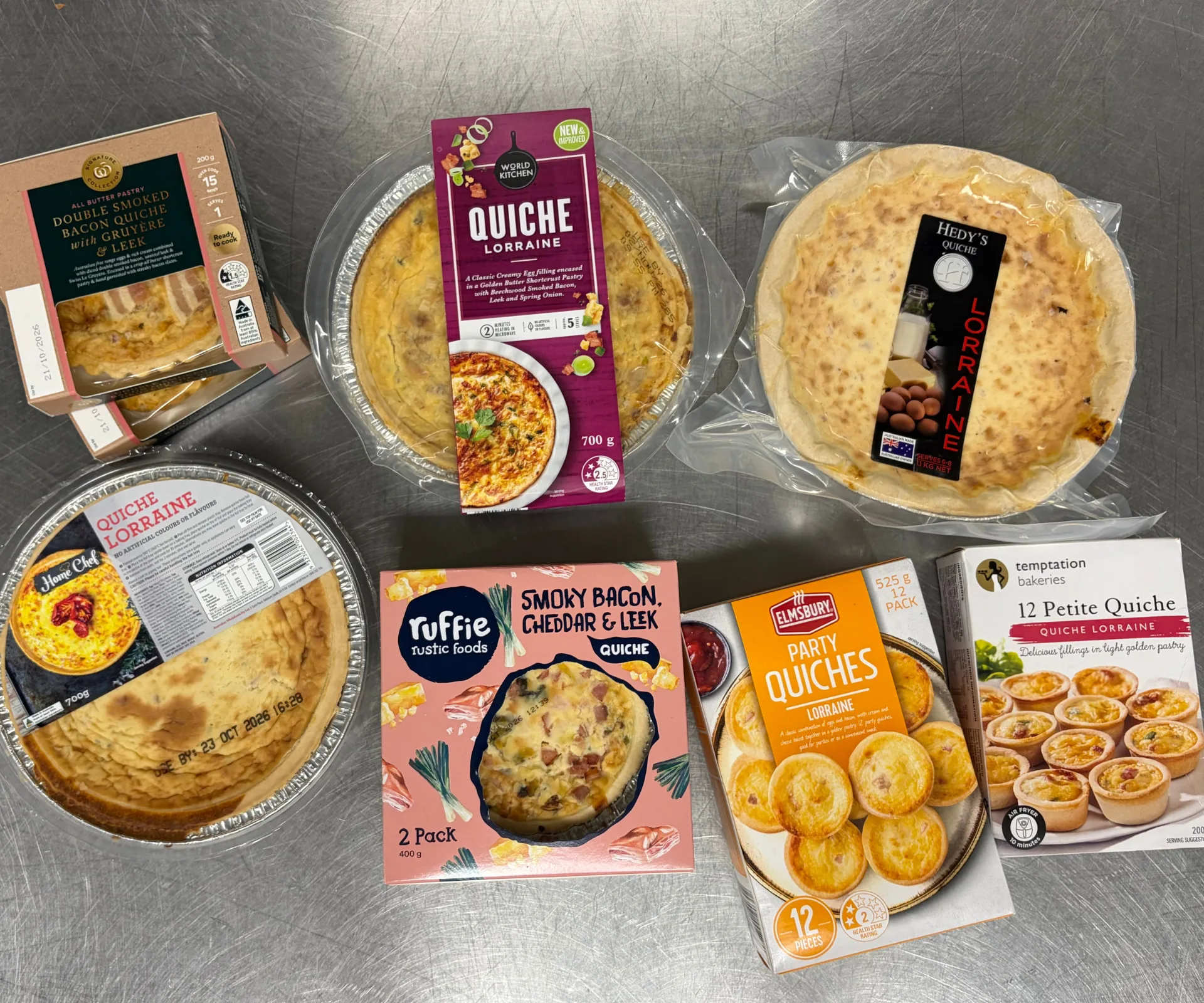 seven supermarket quiches sitting on stainless steel bench, still in their packaging, taken from a birds eye view