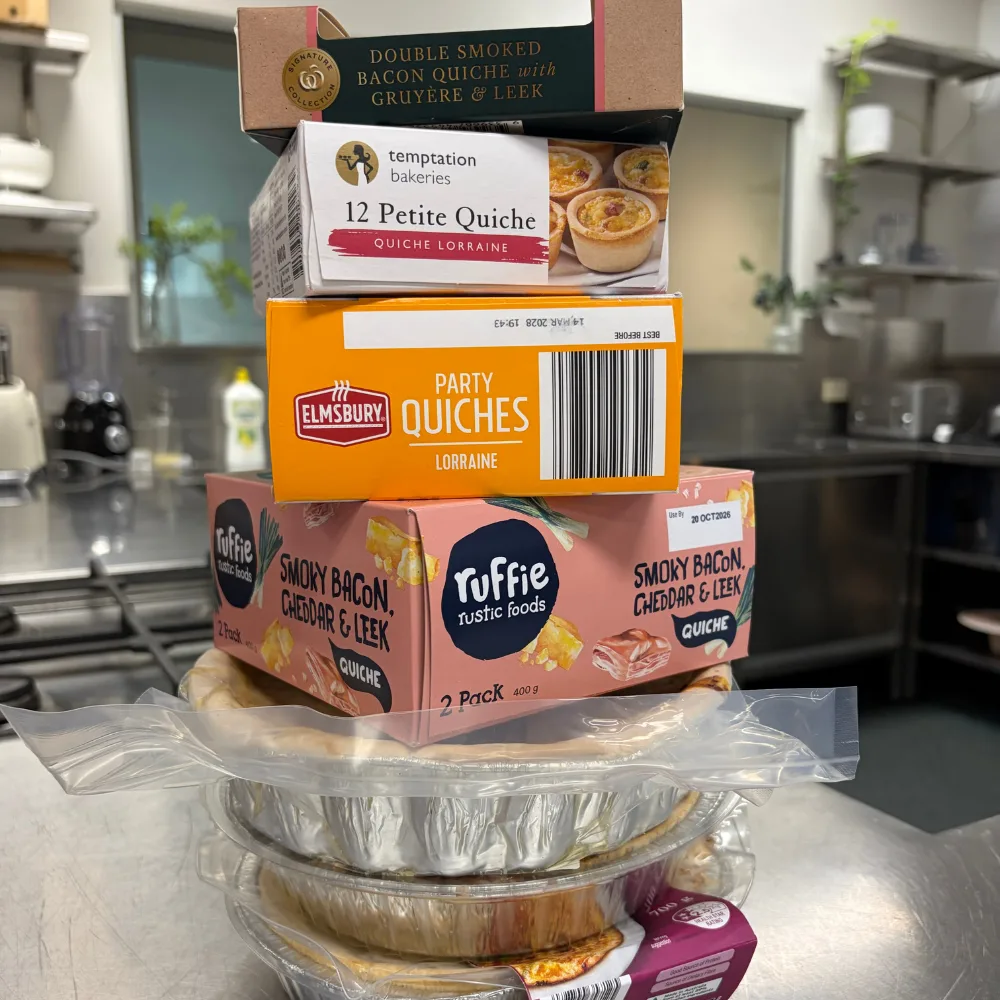 quiches stacked up on top of each other stilll in packaging on stainless steel bench