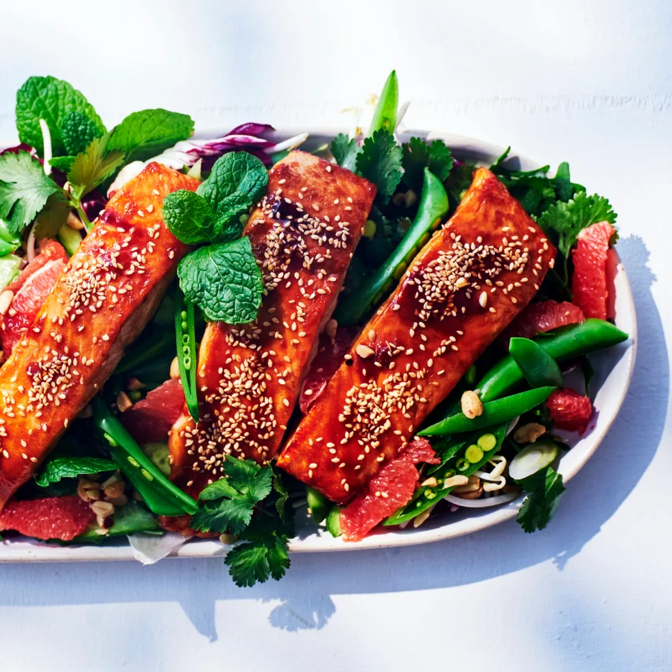 vietnamese baked salmon sitting on long white dish with fresh herbs and sliced snap peas around it. salmon is topped with glaze and white sesame seeds