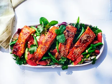 vietnamese baked salmon sitting on long white dish with fresh herbs and sliced snap peas around it. salmon is topped with glaze and white sesame seeds