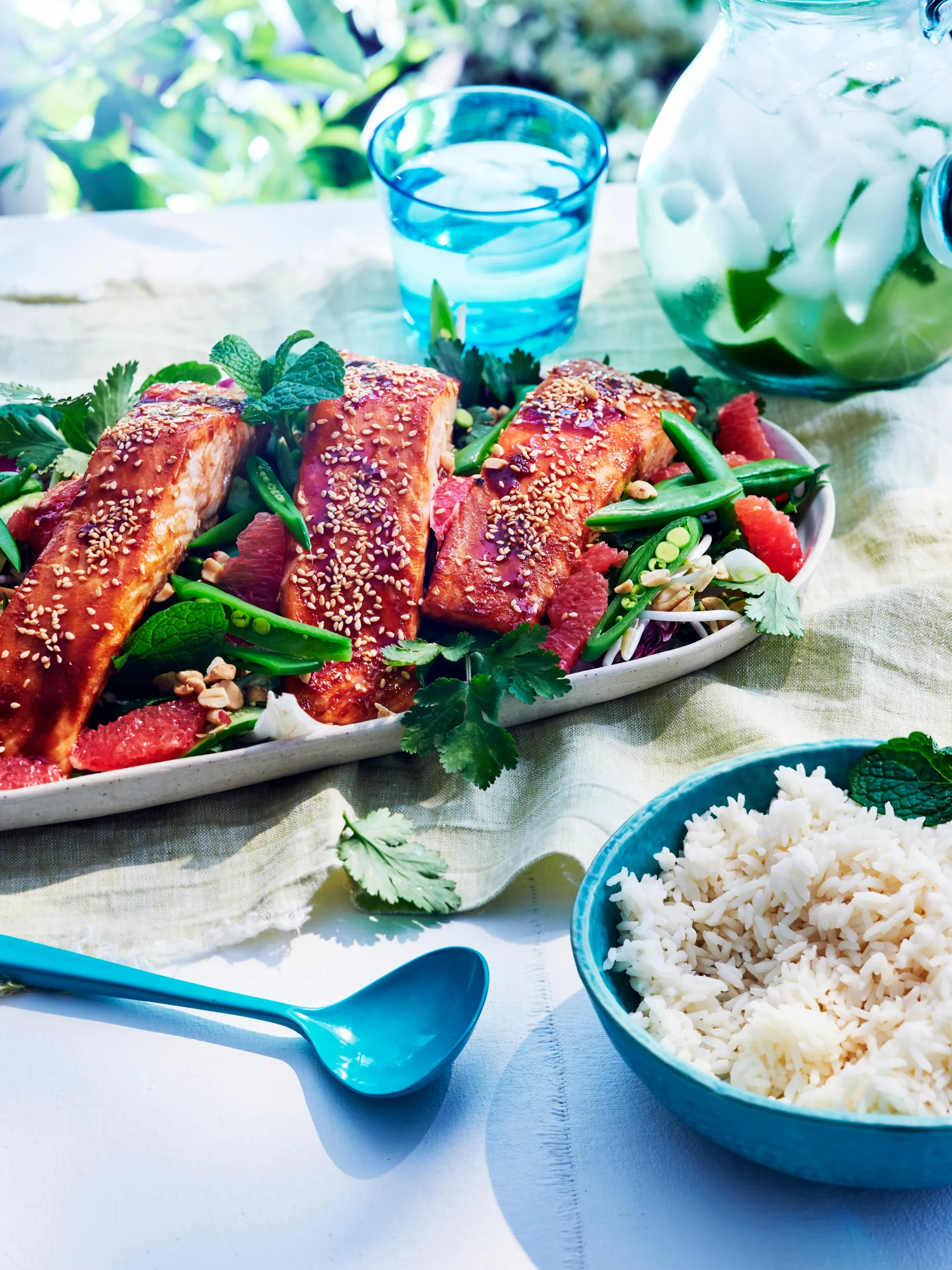vietnamese baked salmon sitting on long white dish with fresh herbs and sliced snap peas around it. salmon is topped with glaze and white sesame seeds