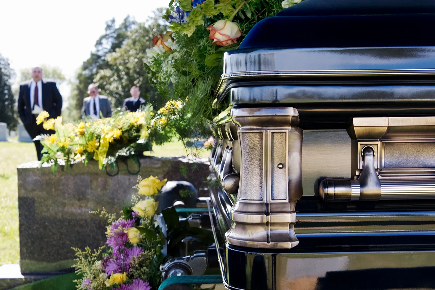 coffin in foreground, mourners in background behind headstone