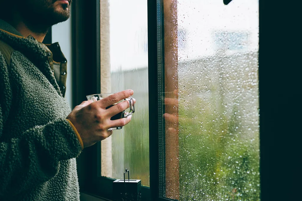 man standing at window with rain with coffee