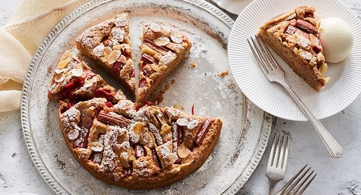 rhubarb cake with flaked almonds and icing sugar on top on rustic metal plate with fork on the side