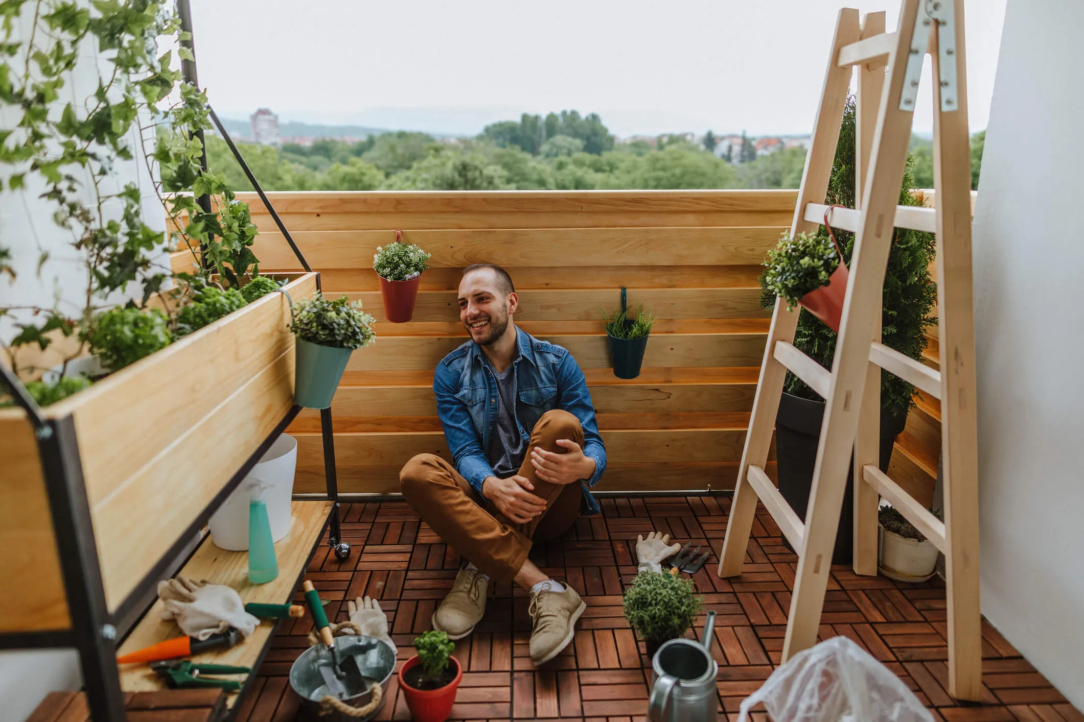 A man sitting on an apartment balcony surrounded by gardening equipment and plants
