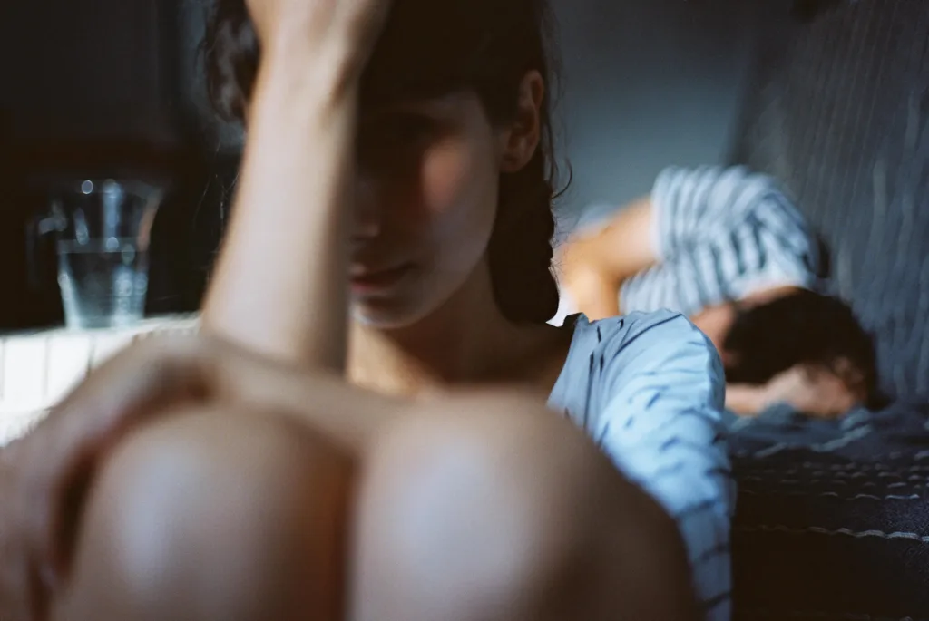 A woman sitting with her head in her hands while her boyfriend lies in the background
