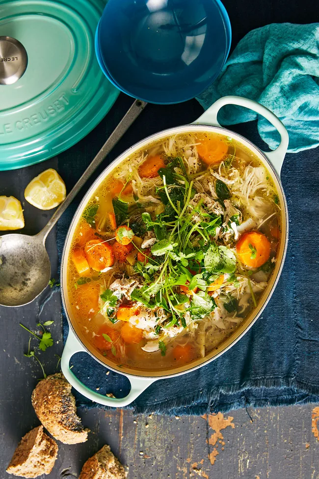Chicken soup with carrot and green vegetables in a white oval pot with handles.