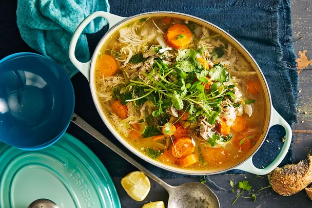 chicken soup with carrot and green herbs in a ceramic white oval dish with a light blue lid next to it.