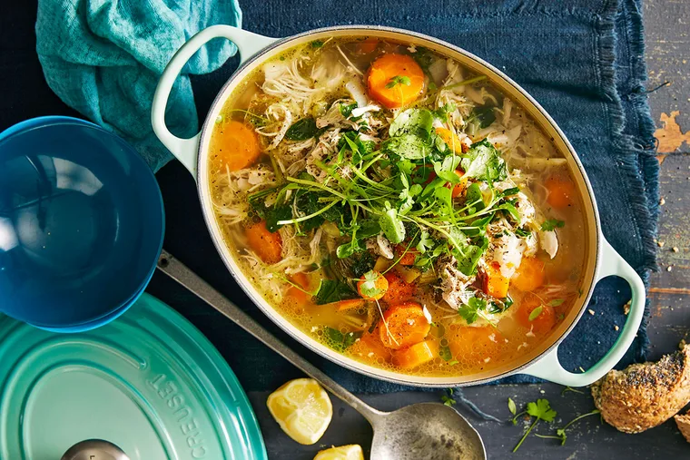 chicken soup with carrot and green herbs in a ceramic white oval dish with a light blue lid next to it.