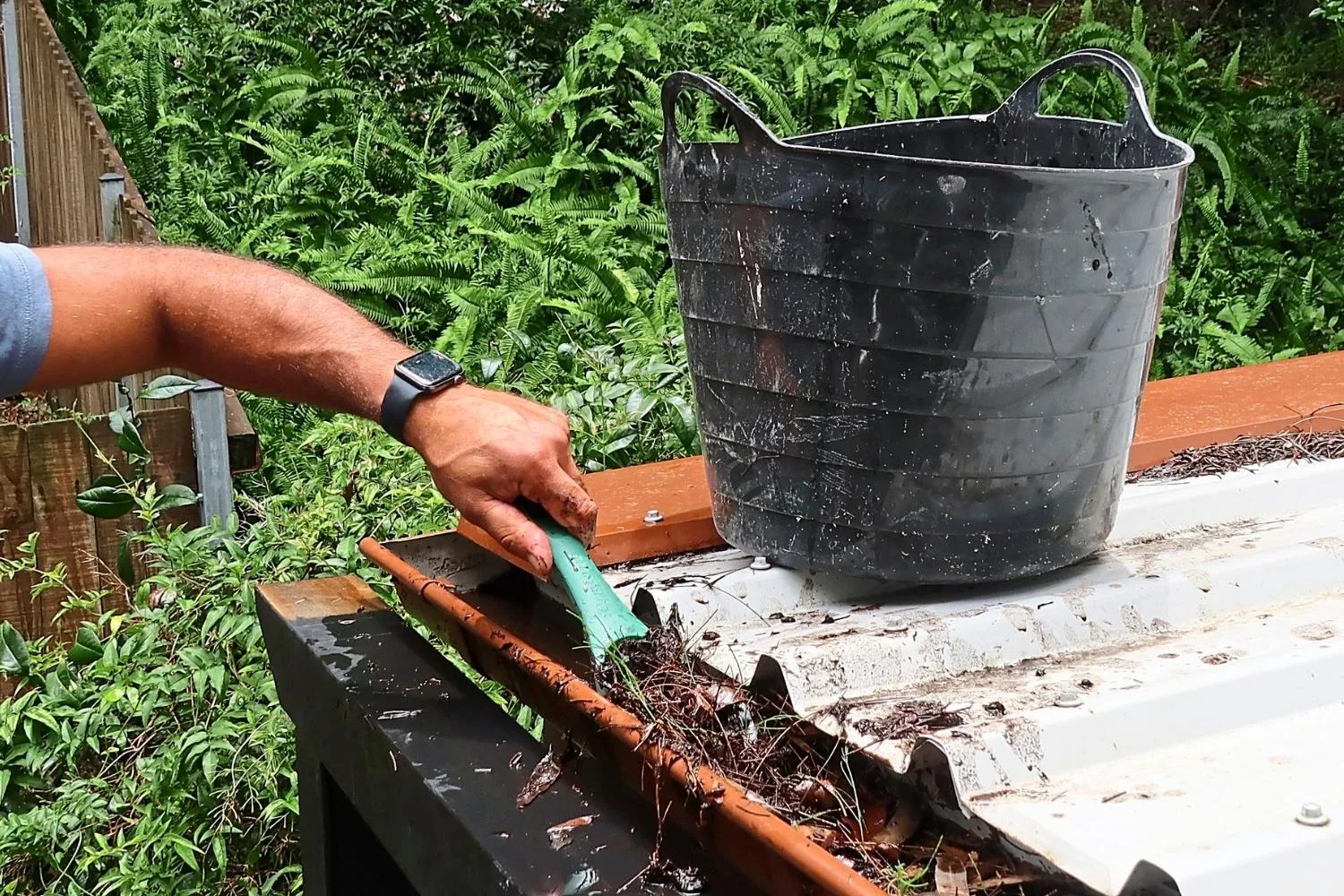 someone clearing out debris from gutter with a scraper and a bin