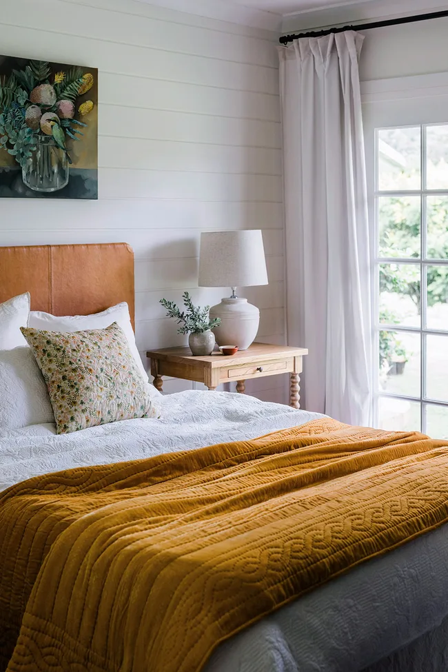 Country bedroom with orange blanket over the bottom of the bed with one throw cushion and a lamp on the bedside table