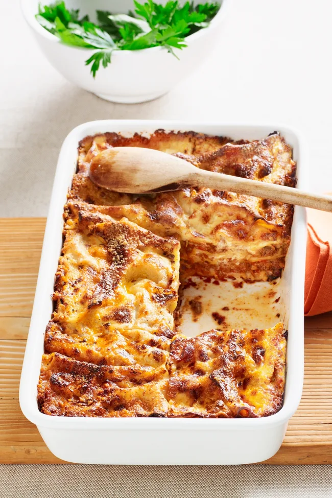 Lasagne dish with one lasagne slice missing in white rectangular tray with wooden spoon on top.