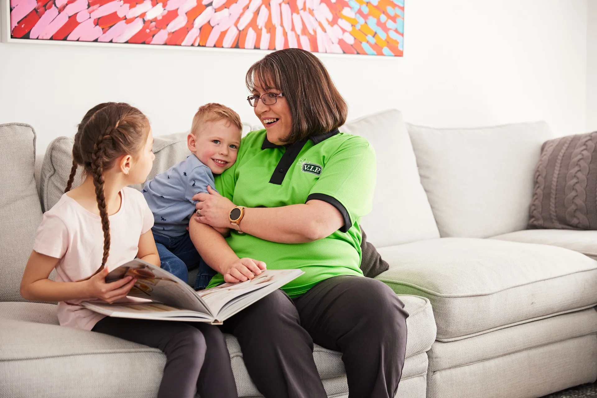 Woman in green shirt sits on sofa with two children, holding a book, smiling and talking.