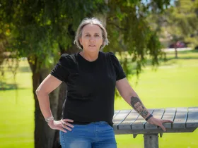 Woman standing outside by a bench in front of a park.