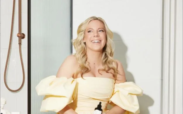 A woman in a light yellow dress sits near a shower holding a bottle, smiling brightly.