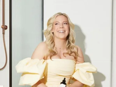 A woman in a light yellow dress sits near a shower holding a bottle, smiling brightly.