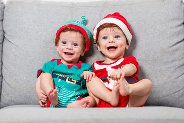 Twins Lucas and Bailey sit side by side on a lounge wearing Christmas outfits. Lucas is dressed as an elf and Bailey is dressed as santa