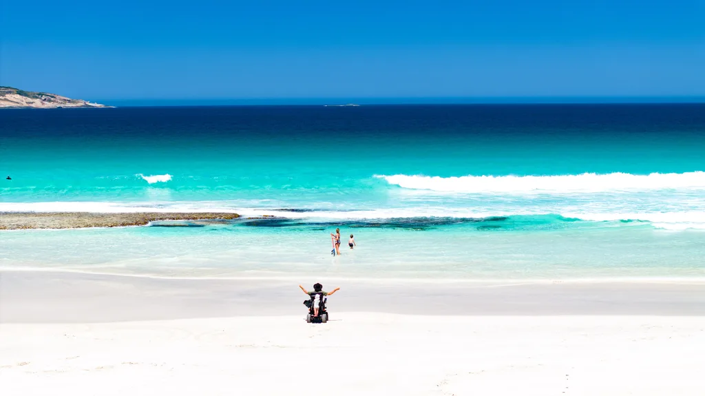 man in wheelchair on white sand looking at aqua blue sea with woman and child in it