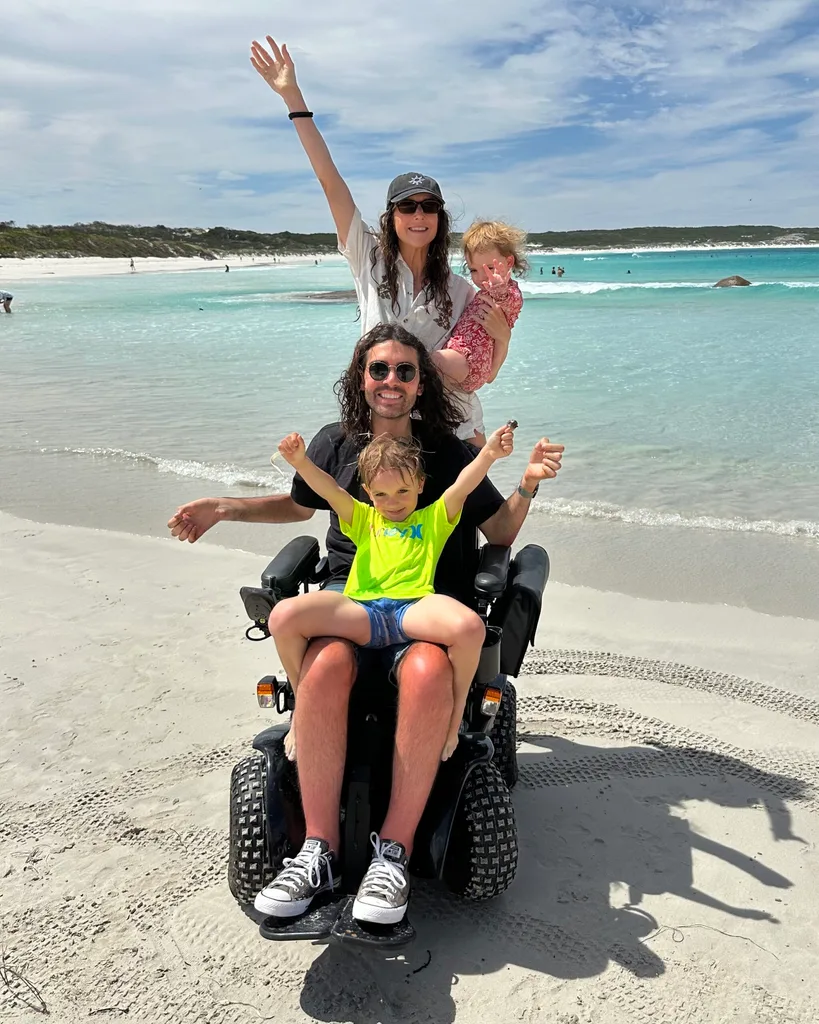 Man in wheelchair with child sat on lap and woman stood behind him holding baby on sand in front of sea