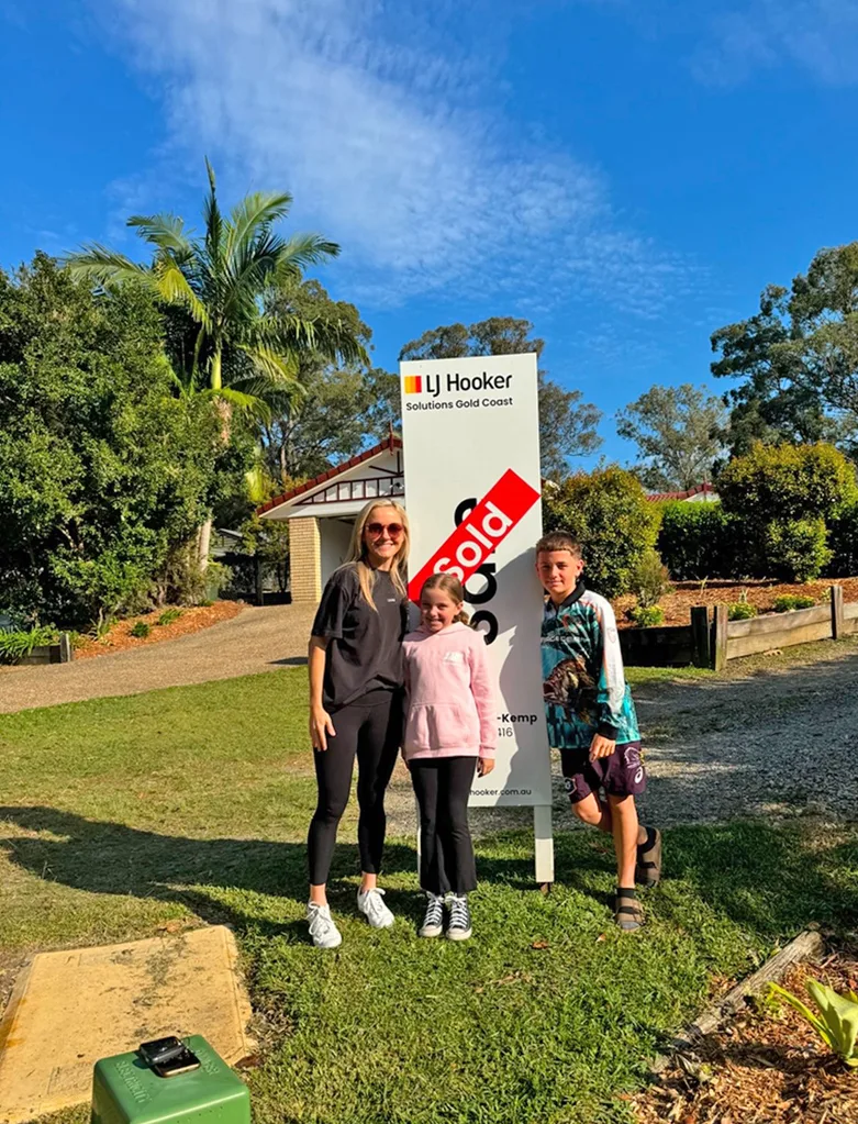 Sarah and her children Lucy and Coby outside in front of the dream house they bought. They are standing with the Sold sign.