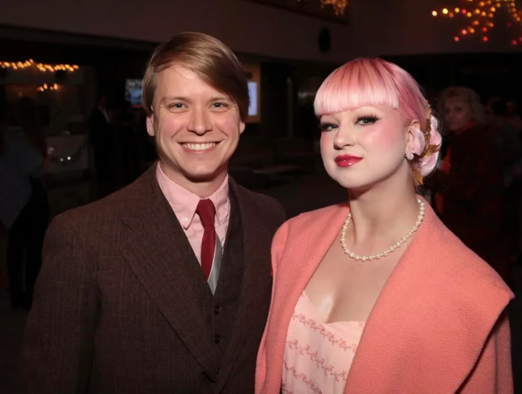 Rochelle wearing a pink wig, pink dress and pink coat, standing next to her husband Matt