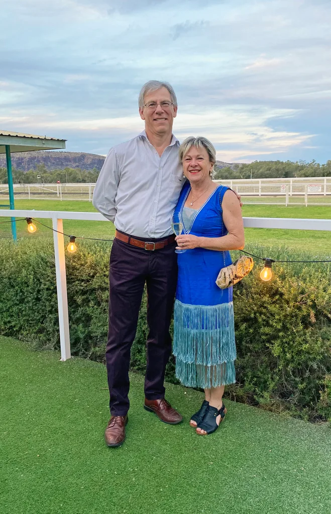 Tanya and her husband Steve smiling for a photo outside. he is wearing a grey button-up shirt, navy pants and a brown belt. She is a wearing a bright blue dress with teal fringe at the bottom.