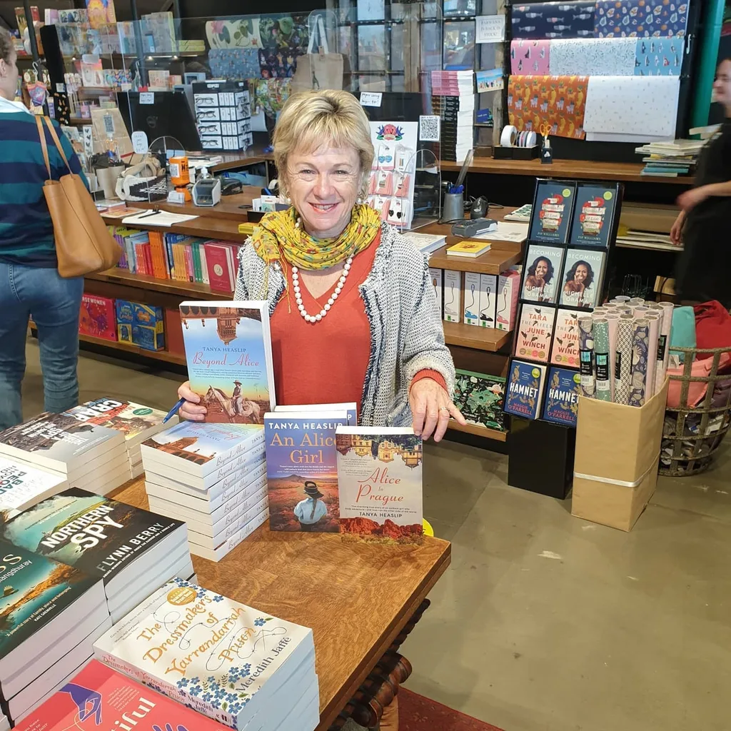 Tanya at a book signing event with all her books. She has short blonde hair and is wearing a coral-coloured top, grey caridgan, yellow floral scarf and a string of white pearls around her neck.