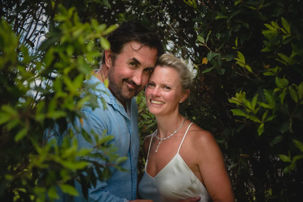 John and Claire at their wedding in 2017. They are smiling together while standing by some trees.