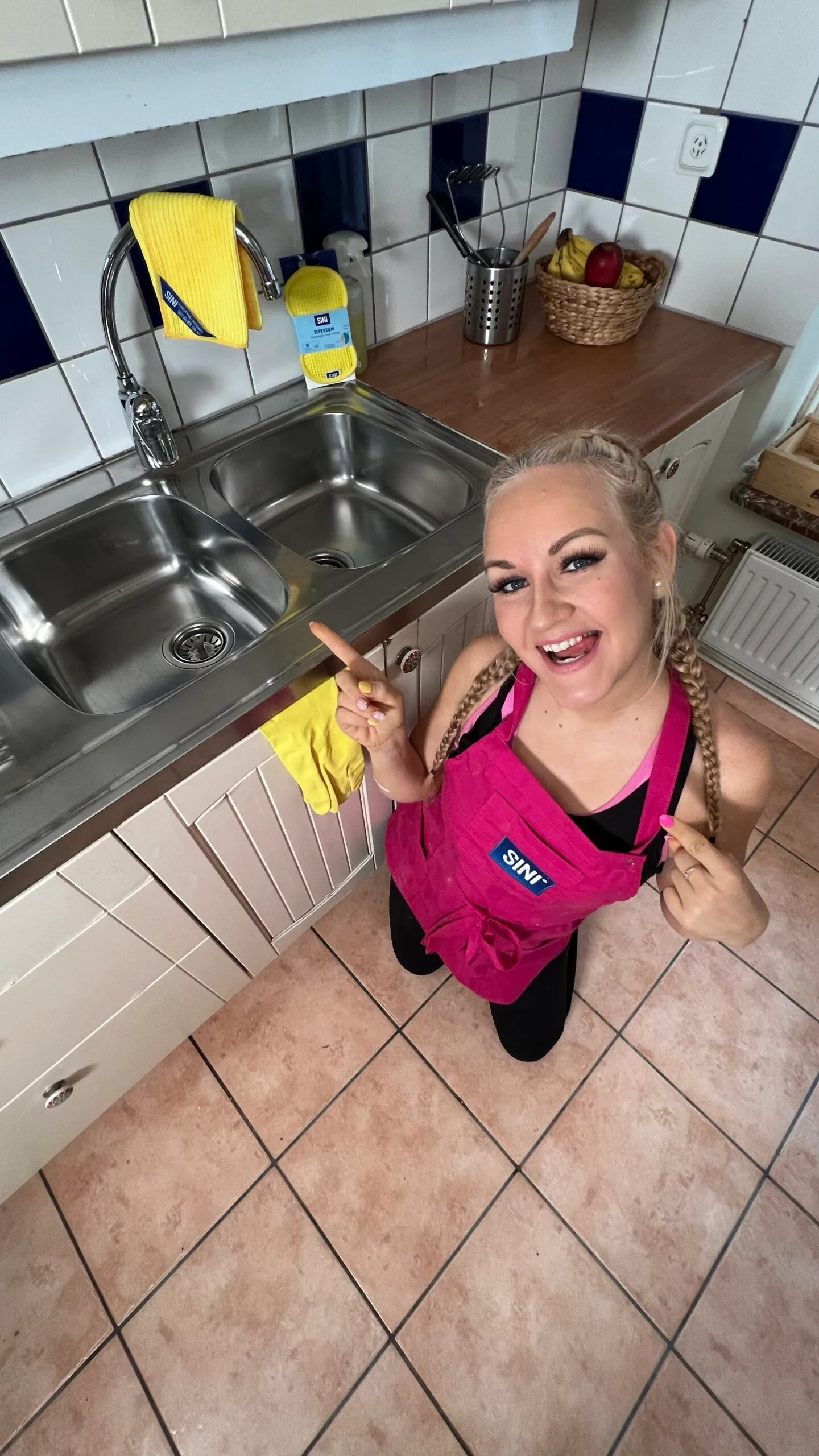 Auri in a clean kitchen. She is pointing at the sink. And she's wearing her  pink apron.