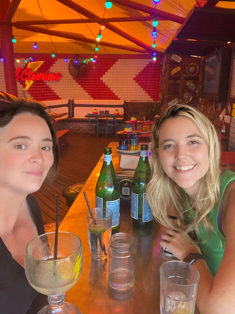 Cassie with a friend at a public venue, sitting at a table with some drinks and smiling at the camera.